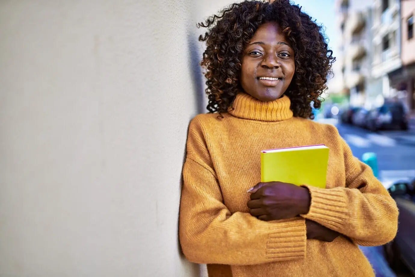 Eine selbstbewusst lächelnde afroamerikanische Frau hält auf der Straße ein Buch.